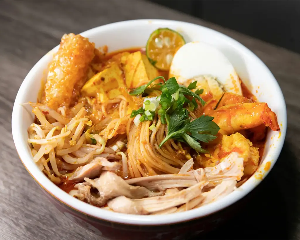 A steaming bowl of Singapore laksa filled with rich, creamy coconut broth, topped with prawns, fish cake slices, tofu puffs, and thick rice noodles, garnished with fresh laksa leaves and a hint of sambal for added depth.
