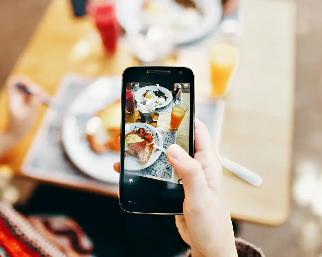 A person’s hand holding a smartphone, carefully framing a beautifully plated dish, ready to capture the perfect food shot at a dining table.