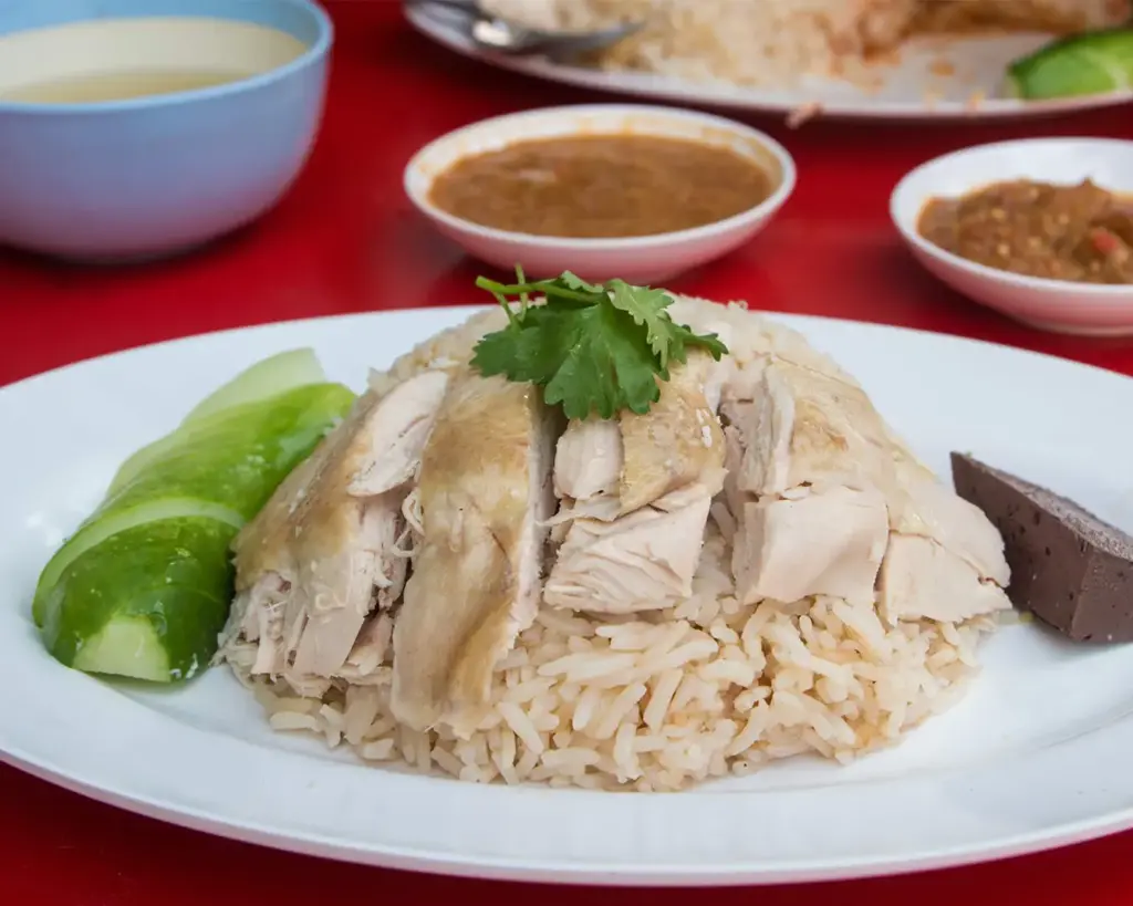 A plate of chicken rice served on a dining table, featuring tender slices of poached chicken arranged neatly over fragrant rice. The dish is accompanied by small bowls of chilli sauce.