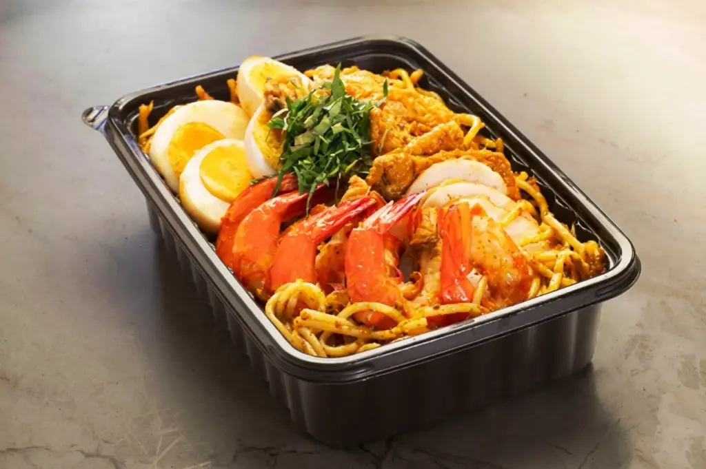 A plastic takeaway container of premium dry laksa from Qi Ji in Singapore, packed with stir-fried laksa noodles, large whole prawns, hard-boiled egg slices, and chopped laksa leaves.