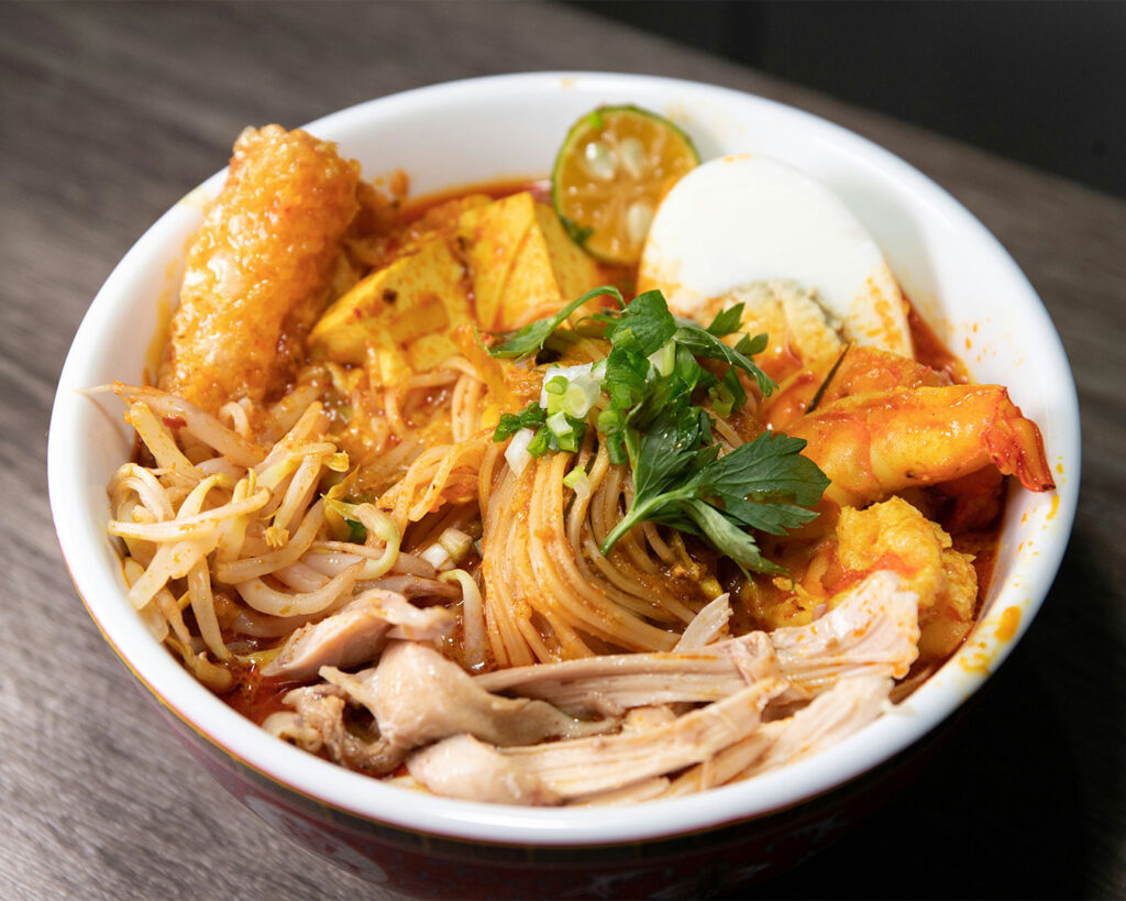 A steaming bowl of laksa with rich coconut curry broth, topped with noodles, prawns, tofu puffs, and fresh herbs, highlighting its bold and aromatic flavours.