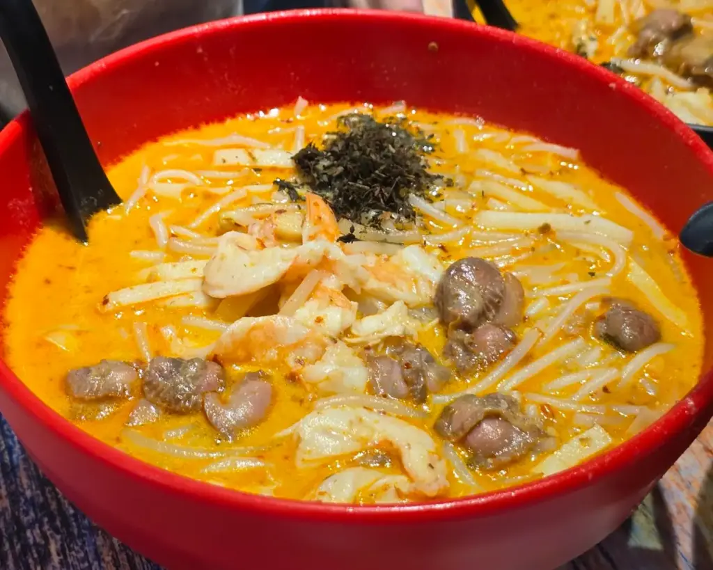 A vibrant bowl of traditional Katong laksa featuring short-cut noodles in a fragrant, spicy coconut gravy, served with prawns, cockles, and fish cake, designed to be eaten with just a spoon for an authentic local experience.