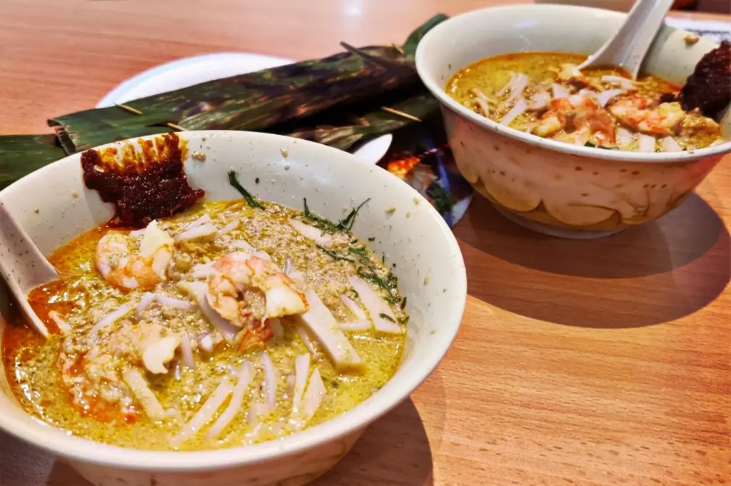 Two bowls of original Katong Laksa from Janggut Laksa in Singapore, served with fresh prawns, thick rice noodles, and a side of otah wrapped in banana leaves.