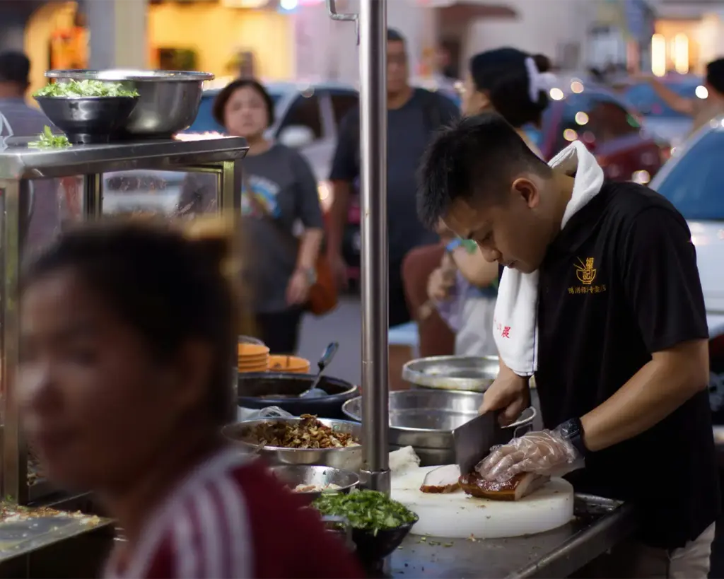 A hawker preparing fresh food, slicing tender roasted chicken with precision at a bustling street food stall, surrounded by vibrant ingredients and cooking tools.