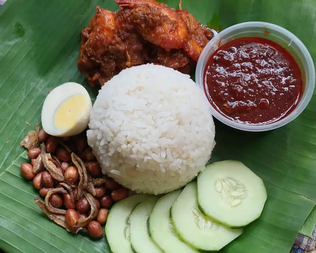 Nasi lemak served on a fresh banana leaf, featuring coconut rice accompanied by sambal, anchovies, peanuts, cucumber, and egg in a traditional Malaysian presentation.