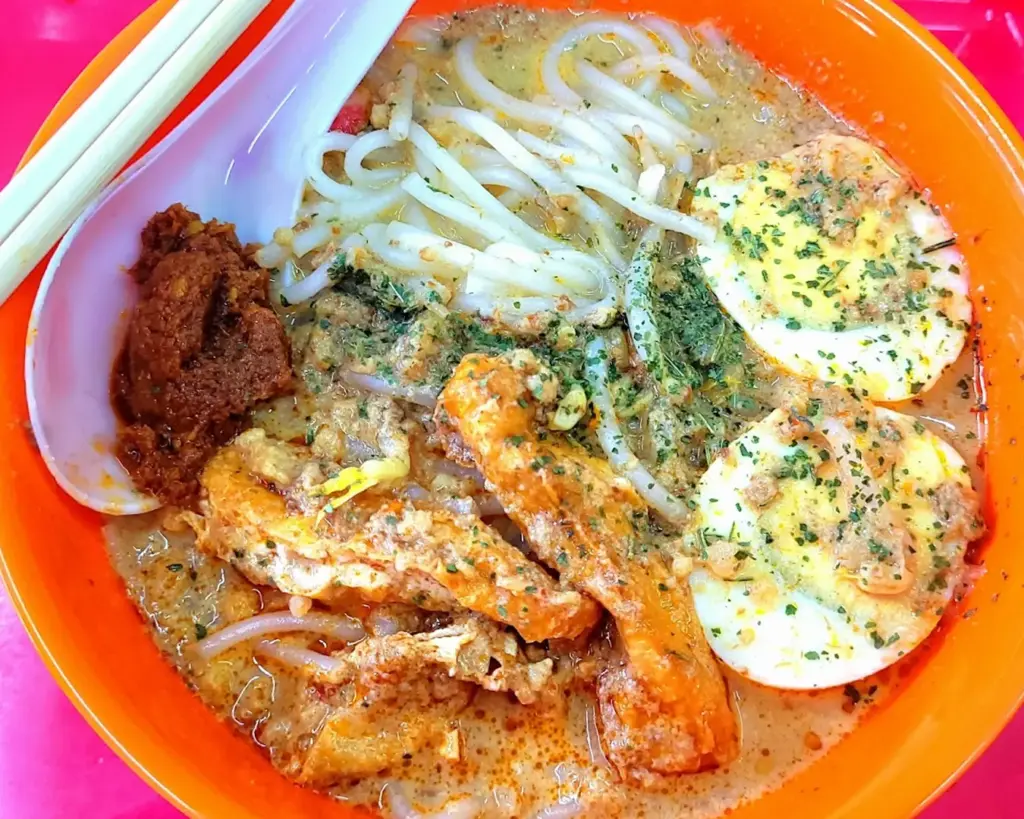 A hearty bowl of Yishun-style laksa with a slightly thicker, robust broth, packed with prawns, tofu puffs, and noodles, finished with a generous spoonful of sambal chilli that adds a bold and spicy kick.
