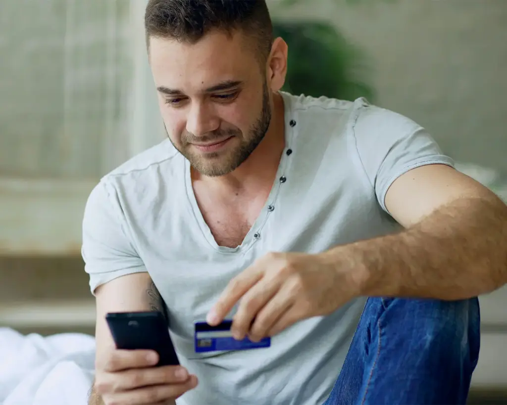 A man using his smartphone to complete a digital payment while holding a bank card in his other hand. The moment captures the ease of mobile payment