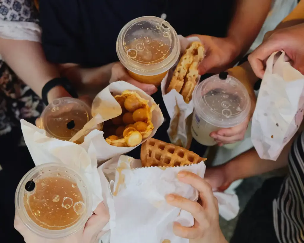 Hands holding a freshly made waffle wrap and other hands holding clear takeaway cups filled with iced beverages