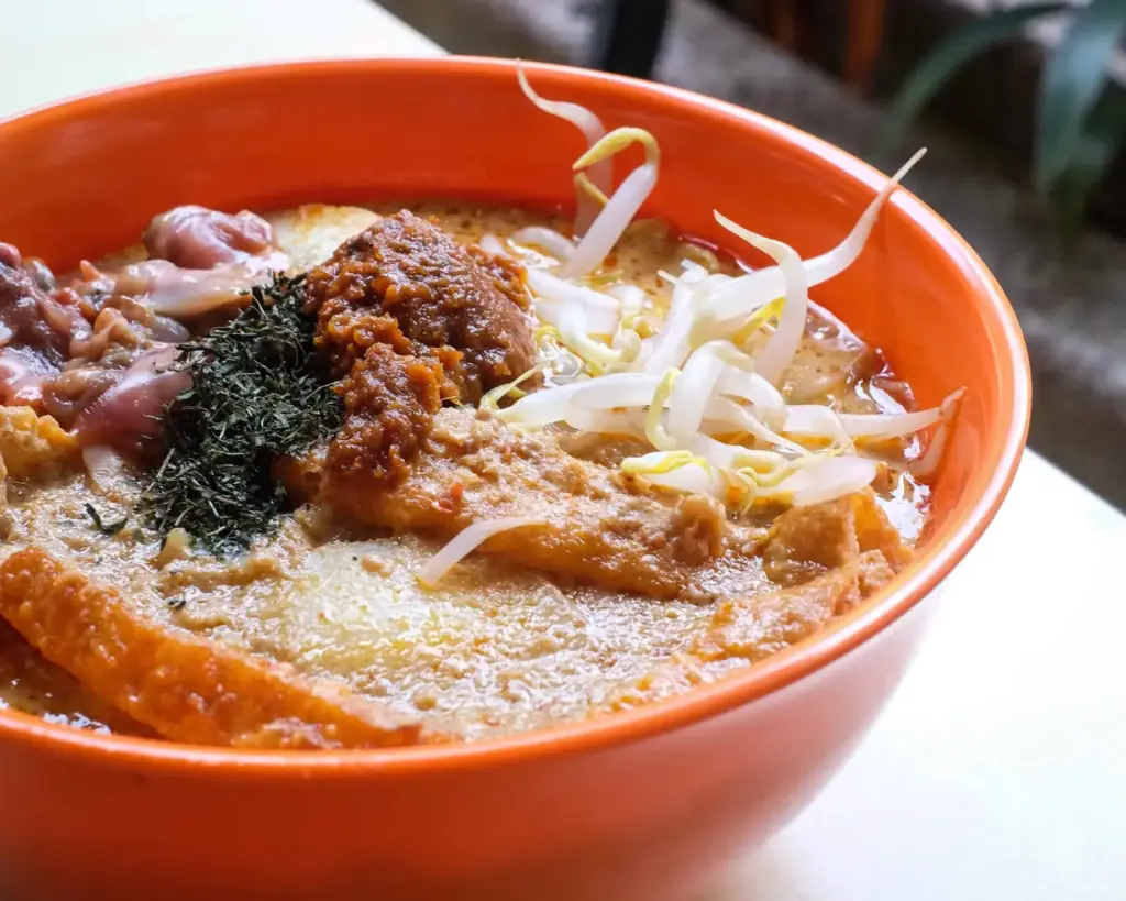 A comforting bowl of Singapore laksa showcasing a golden, aromatic coconut soup base, layered with seafood, noodles, and herbs, creating a rich and satisfying dish that reflects the city’s diverse flavours.
