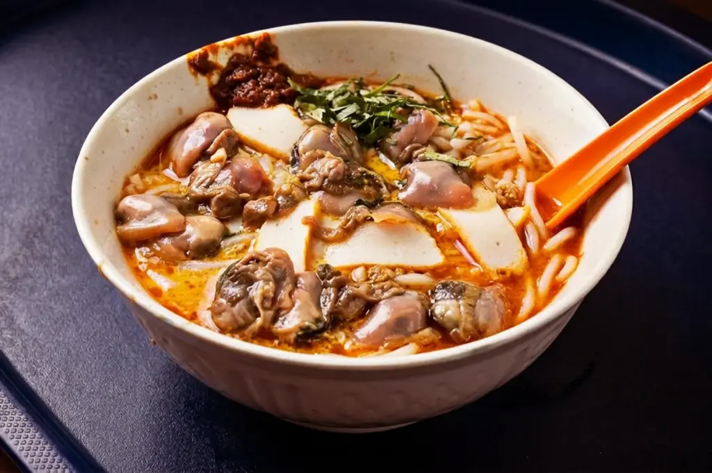 A bowl of traditional Sungei Road Laksa in Singapore heavily topped with fresh cockles, sliced fishcakes, and sambal chili paste.