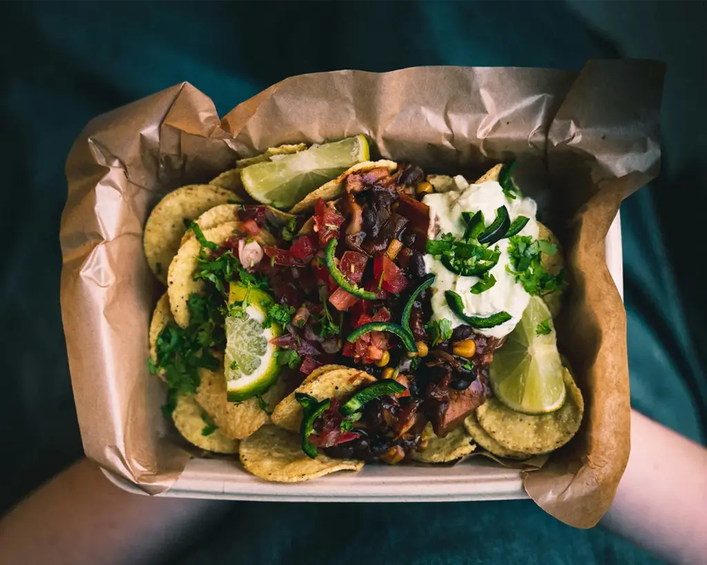 A hand lifting a generous serving of street-style nachos loaded with cheese, salsa, and other colourful toppings, ready to enjoy at an outdoor food market.