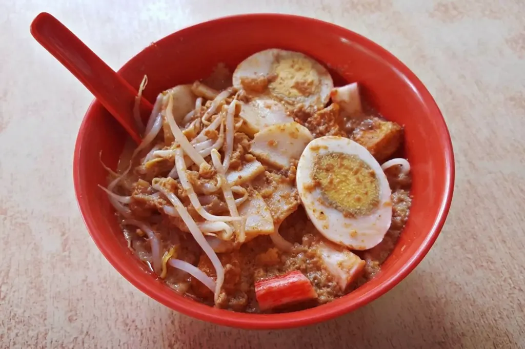 A vibrant red bowl of famous 928 Yishun Laksa in Singapore, loaded with thick rice noodles, hard-boiled eggs, bean sprouts, and fishcake slices in a rich broth.