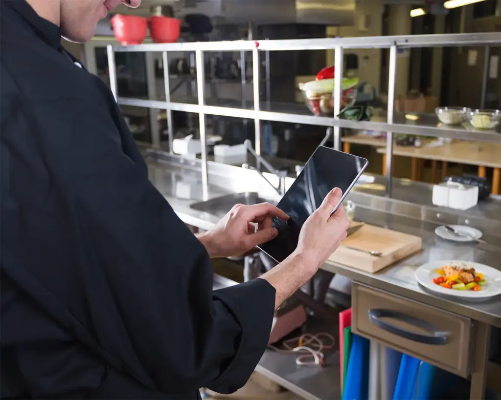 A chef standing in a kitchen while checking incoming orders on a tablet device. The screen displays order details, reflecting how digital tools help kitchens manage and organise food preparation.