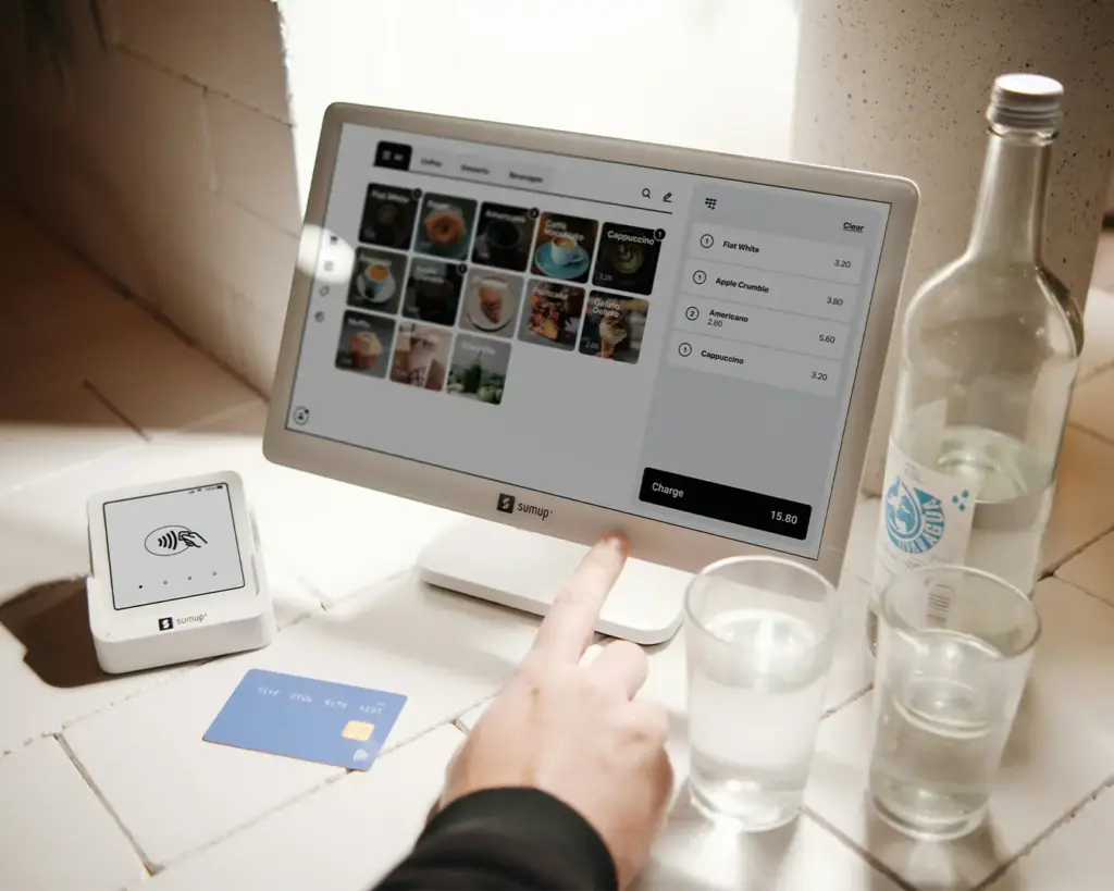 A hand interacting with a touchscreen digital menu at a café, selecting food or drink options. The interface represents a modern ordering experience where customers browse and choose items digitally.