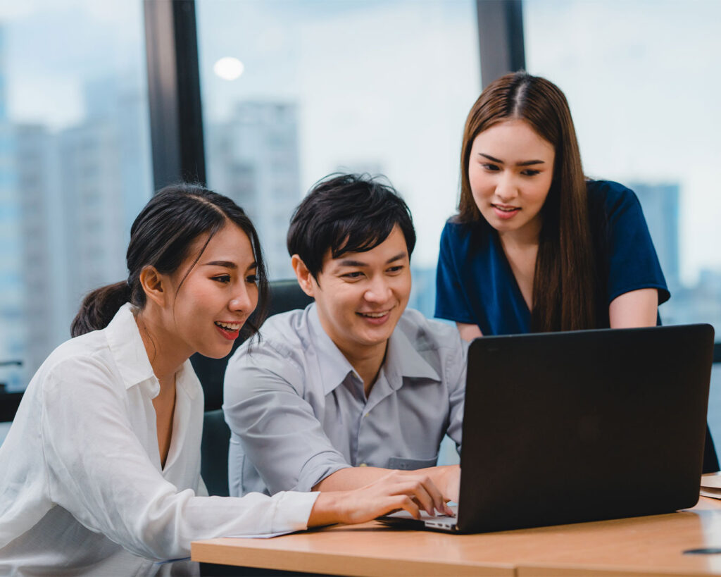 A small team gathered around a laptop, discussing ideas and pointing at the screen during a collaborative meeting in a modern workspace.