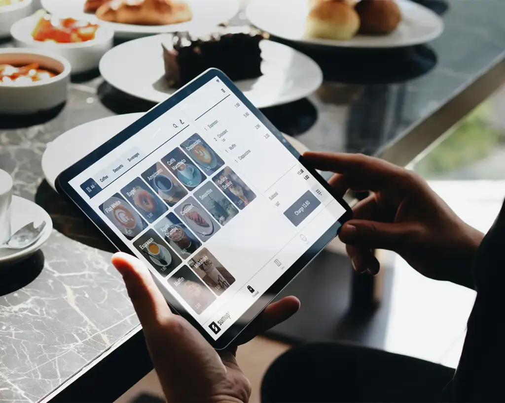 A person seated at a café table holding a tablet to view the menu while eating. The scene highlights the convenience of browsing food options digitally during a relaxed dining experience.