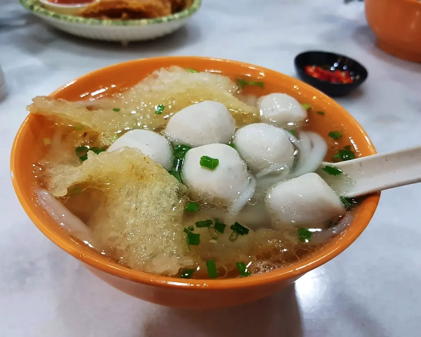 A vibrant orange bowl filled with fishball soup, featuring white fishballs, fried fish skin, noodles, and chopped green onions, next to a black dish of chili.