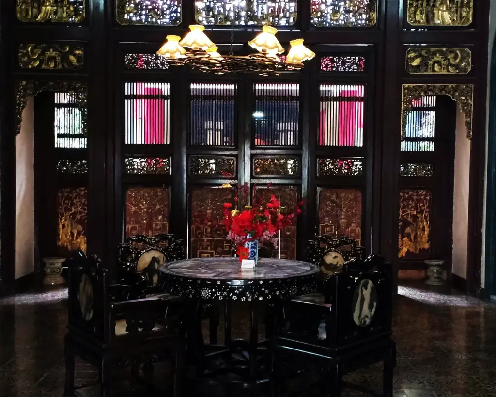 Elegant dining room with ornate dark wood decor and intricate carvings. Round table with a floral centerpiece under a warm chandelier, creating a cozy, historic ambiance.