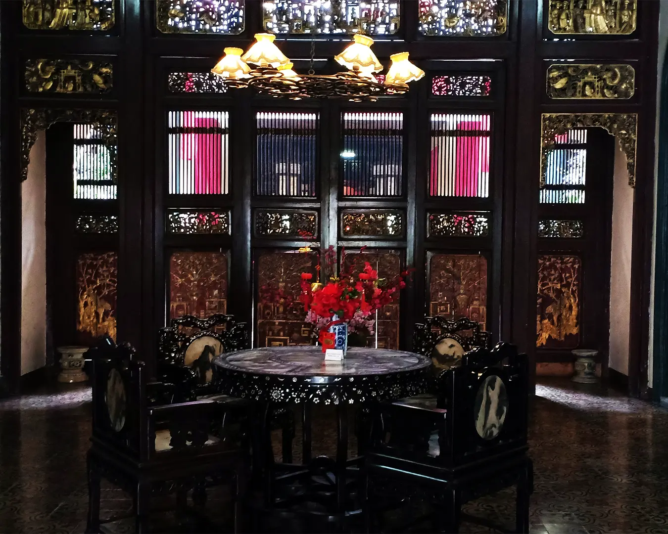 Elegant dining room with ornate dark wood decor and intricate carvings. Round table with a floral centerpiece under a warm chandelier, creating a cozy, historic ambiance.