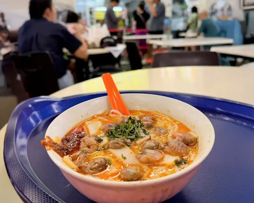 Bowl of spicy noodle soup with clams and herbs, served with an orange spoon on a blue tray in a busy food court.