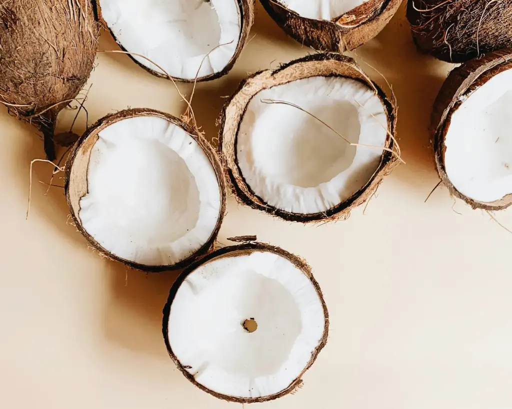 Halved coconuts with rough brown shells and smooth white flesh are arranged on a light surface. The image conveys a fresh, natural feel.
