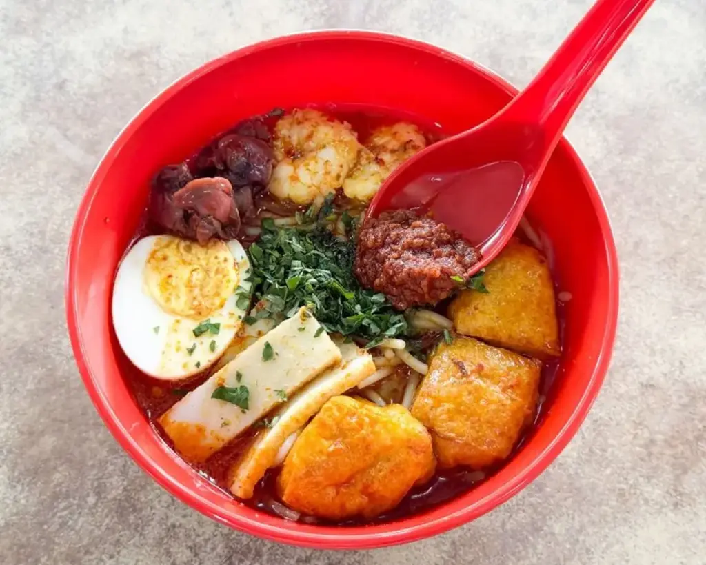 A vibrant red bowl of spicy noodle soup topped with shrimp, tofu, boiled egg, greens, and a dollop of chili paste, with a red spoon on the side.