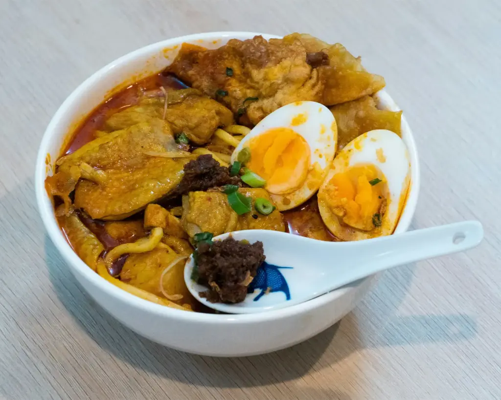 Bowl of spicy noodle soup with sliced boiled egg, crispy meat, and spring onions. The rich broth conveys a warm, savory atmosphere.
