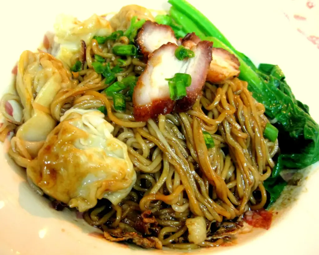A plate of kampua noodles tossed in fragrant oil and seasoning, topped with slices of char siu and minced pork, reflecting the dry-style noodle dish known for its light yet flavourful profile.