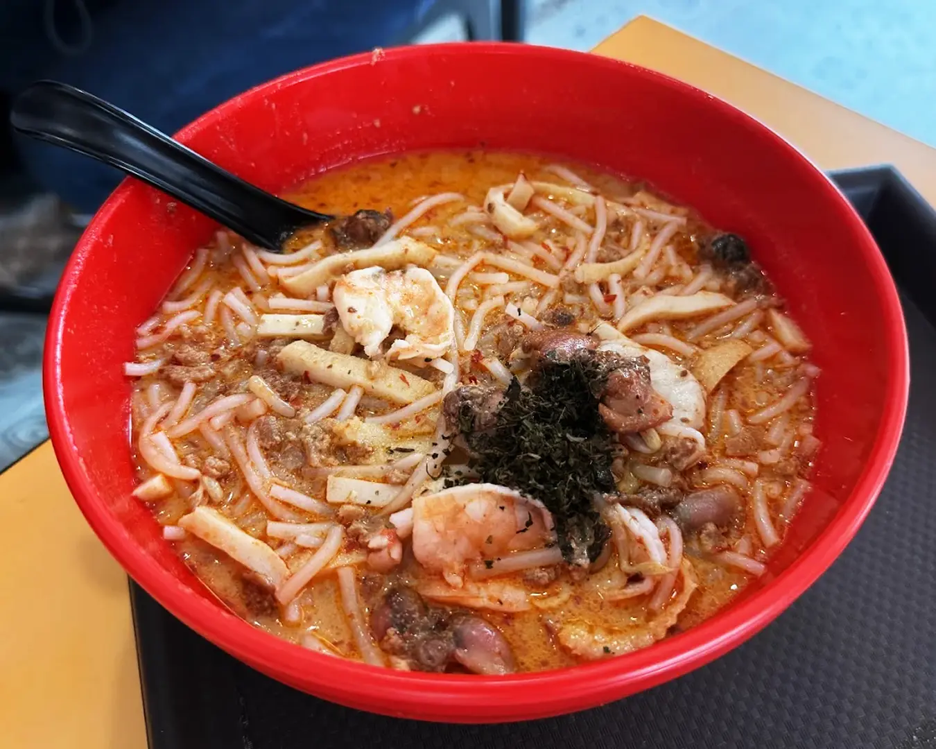 A vibrant red bowl of spicy laksa is filled with noodles, prawns, tofu, and topped with herbs. A black spoon rests on the edge, ready to enjoy.