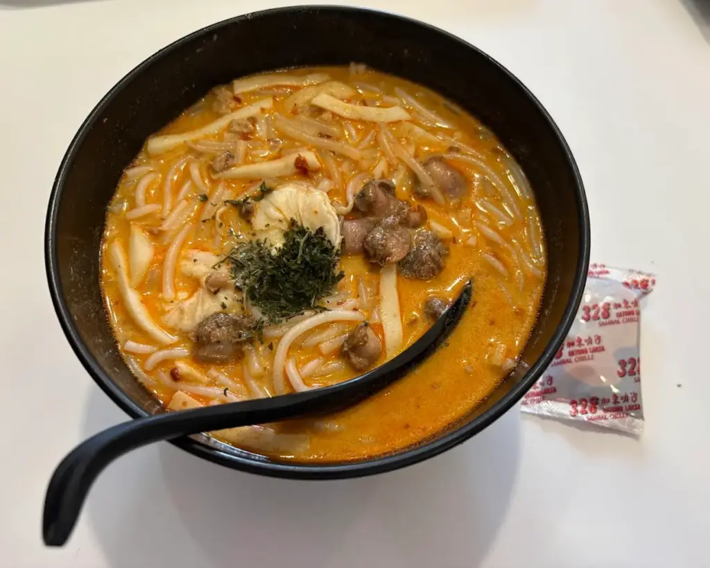 A bowl of spicy noodle soup with chicken, garnished with herbs, on a white surface. A black spoon and a packet of chili sauce are beside it.