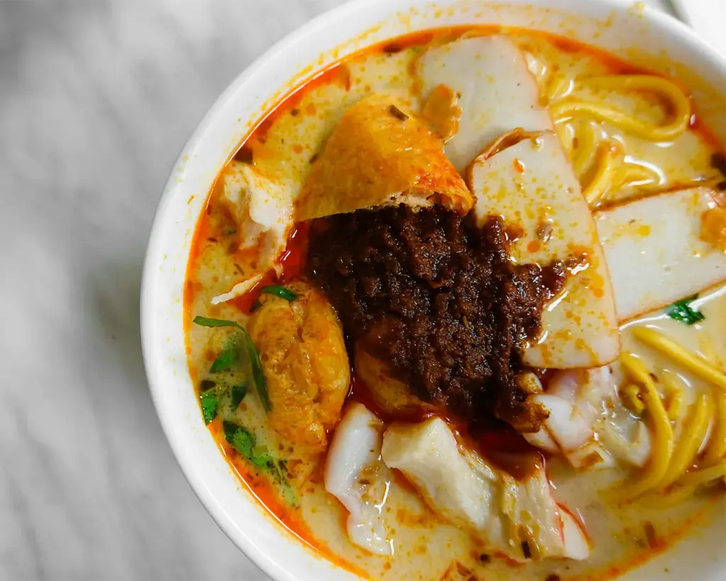 A vibrant bowl of curry laksa with noodles, tofu, fish cakes, and a dollop of spicy sambal. The rich broth glows with warm, enticing tones.
