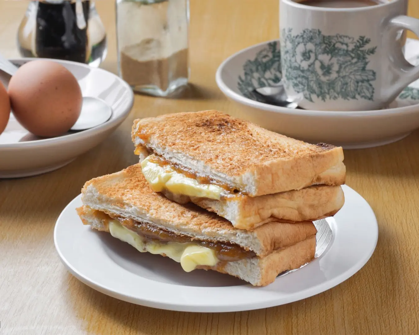 Toasted sandwiches with butter and kaya spread on a white plate, accompanied by two boiled eggs and a cup of black coffee on a wooden table.