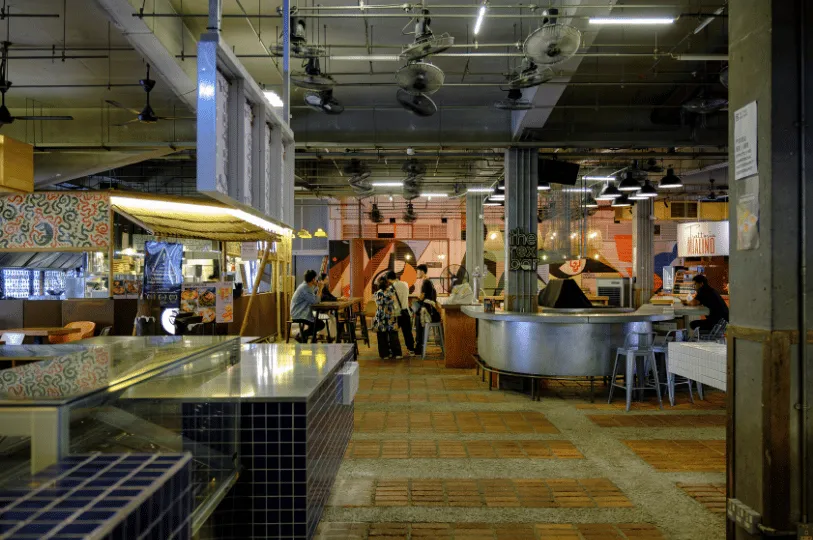 A spacious, modern hawker centre with industrial decor, overhead fans, and plenty of empty tables and counter seating, highlighting a quieter environment with easy seating choices.