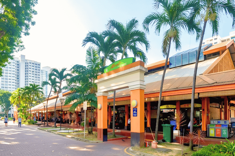Bright exterior shot of Maxwell Food Centre's main entrance on a sunny day, featuring its iconic orange arched gateway, green palm trees, and pedestrians walking outside.