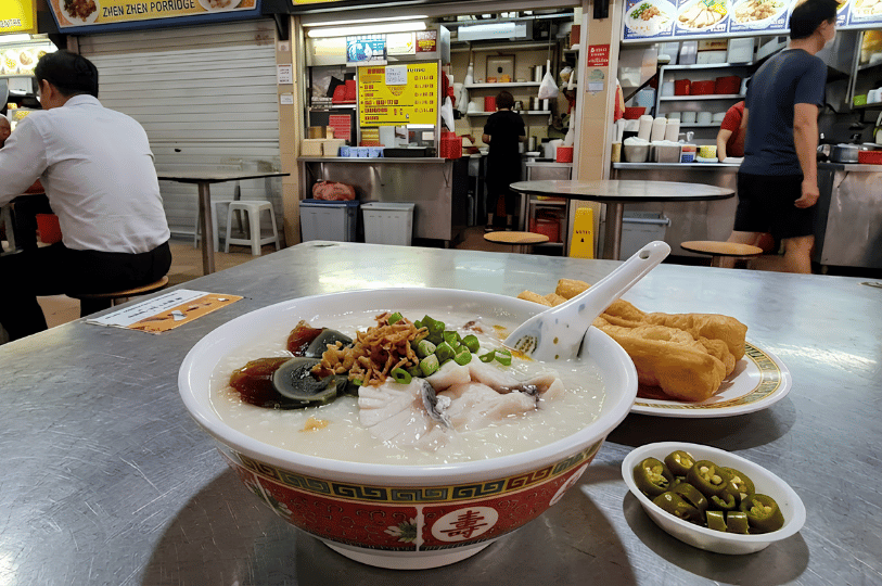 A close-up of a large, comforting bowl of thick congee topped with sliced fish, century egg, and fresh garnishes, accompanied by a plate of crispy fried dough sticks (youtiao) at a hawker centre table.