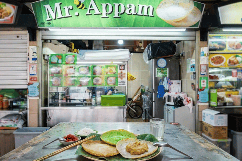 A fresh plate of crispy South Indian appam pancakes served on a banana leaf with grated coconut and bright orange sugar, sitting on a table directly in front of the brightly lit Mr. Appam stall.