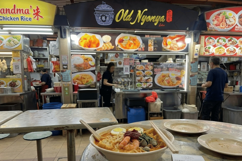 A steaming bowl of spicy laksa loaded with thick noodles, prawns, cockles, tofu puffs, and a dollop of chili paste on a metal table in front of the Old Nyonya hawker stall.