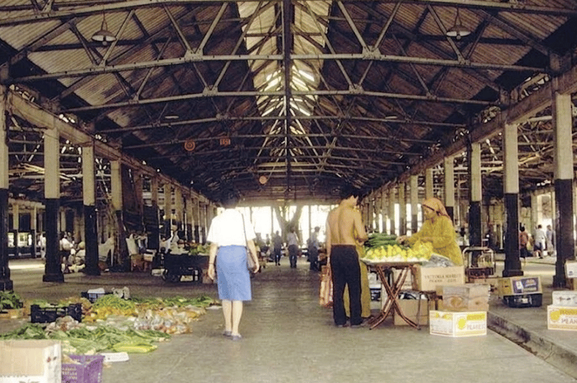 A vintage photograph showing the historic interior of the Maxwell building when it operated as a wet market, featuring vendors selling fresh produce and vegetables on the floor beneath the high metal rafters.
