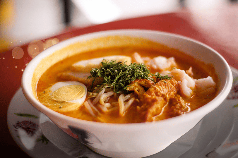 A close-up of a steaming bowl of Singapore laksa, drawing focus to the finely chopped green laksa leaves (daun kesum) garnished on top and the porous, sponge-like tofu puffs (tau pok) soaking in the rich, orange coconut curry broth.