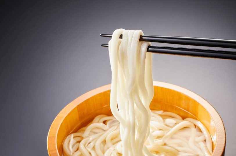 A tight close-up of thick, smooth white noodles being lifted by black chopsticks from a clear broth, highlighting the firm, springy texture that provides the essential foundational bite for a heavy, rich soup.