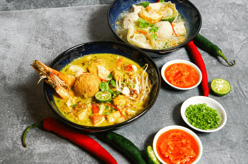 A top-down view of a vibrant bowl of prawn laksa surrounded by its essential, but often overlooked, side condiments: individual small bowls containing bright red sambal chili, freshly chopped green scallions, and halves of fresh calamansi lime for necessary acidity.