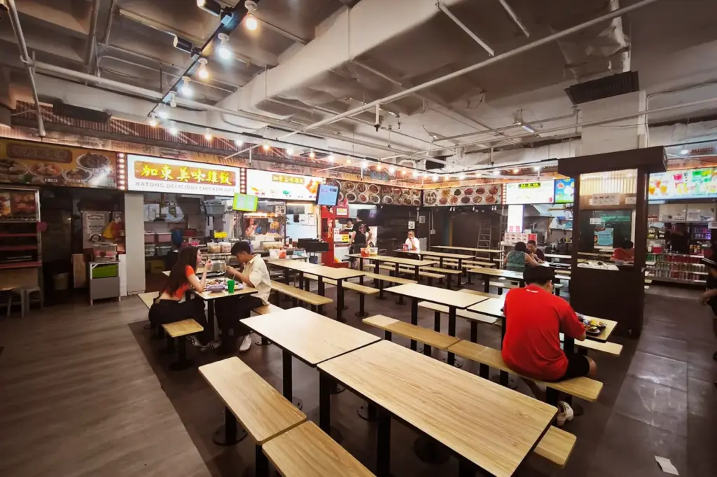 Indoor food court in Singapore featuring Katong Delicious Chicken Rice stall with customers eating at wooden tables.