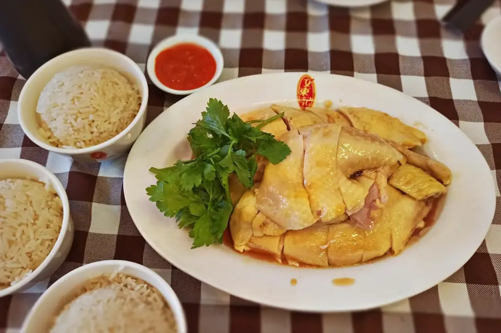 A platter of sliced Hainanese chicken garnished with cilantro, served with bowls of seasoned rice and chili sauce on a checkered tablecloth at Boon Tong Kee restaurant in Singapore.