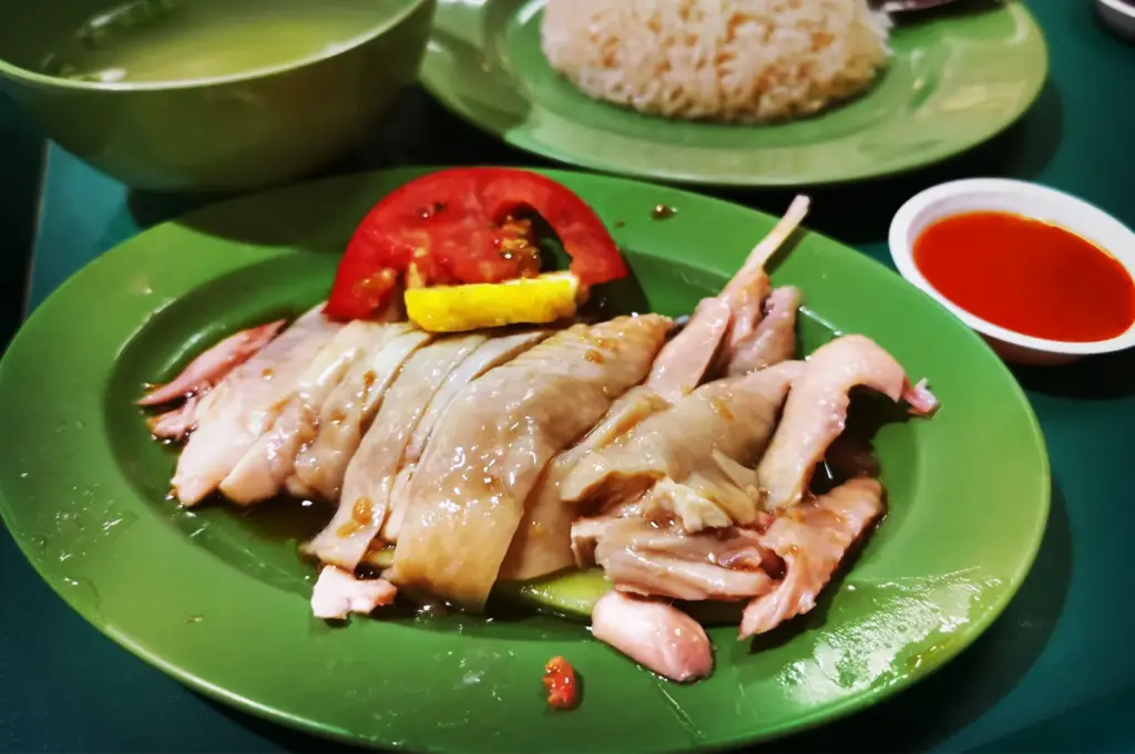 A plate of authentic Xing Yun Hainanese Boneless Chicken Rice in Singapore, served alongside a bowl of seasoned rice, chicken soup, and red chili dipping sauce.
