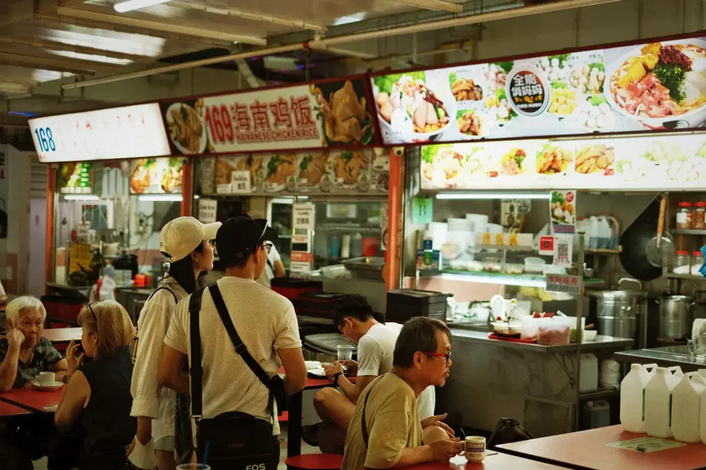 Customers ordering at a Hainanese Chicken Rice stall in a busy Singapore hawker center with people dining at tables.