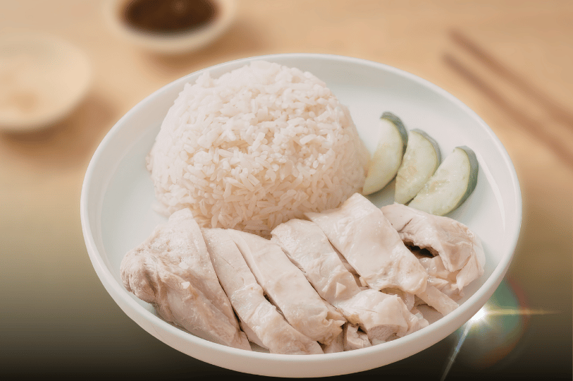 A close-up of a classic plate of Hainanese chicken rice, featuring neatly sliced, smooth poached chicken next to a dome of fragrant rice and fresh cucumber slices.
