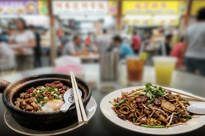 A close-up of two popular local dishes: a steaming dark claypot rice topped with an egg, chicken, and sausage, next to a plate of savory, stir-fried Char Kway Teow noodles. The background features a deeply blurred crowd inside a food centre.