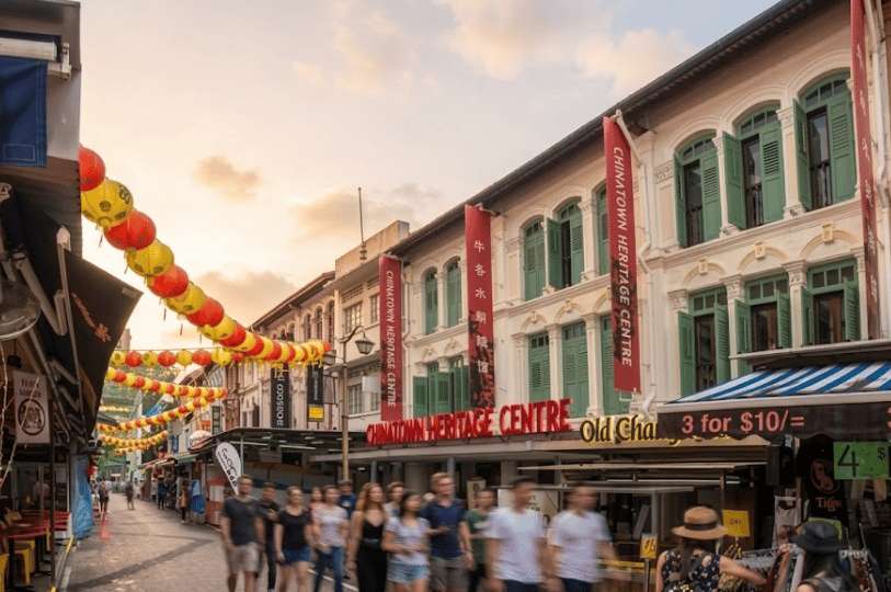 A warm, daytime view of a bustling street in Chinatown. Historic shophouses line the street, featuring bright red banners for the Chinatown Heritage Centre, while strings of lanterns hang overhead. The walking crowd is slightly blurred to convey motion.