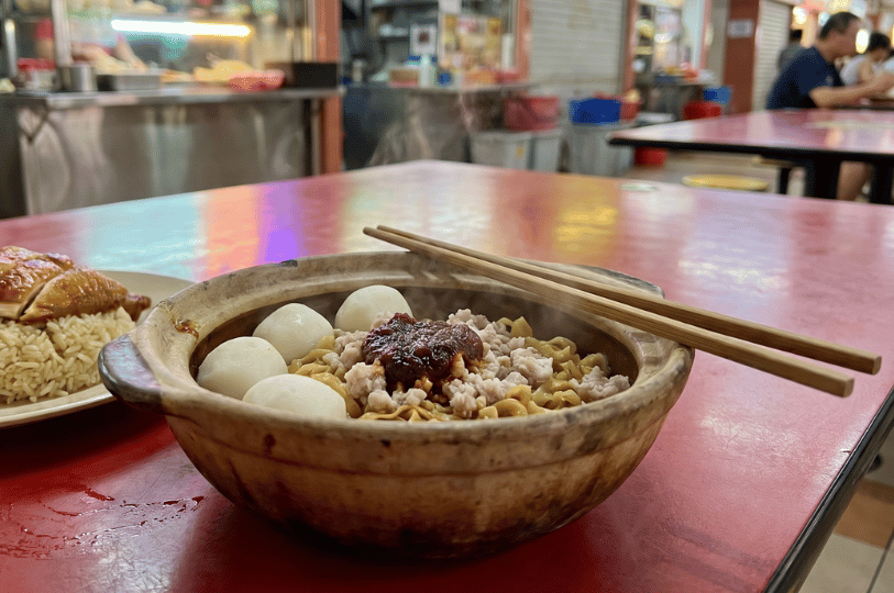 A steaming claypot bowl of noodles topped with four white fishballs, minced meat, and a dollop of dark red chili paste, sitting on a bright red table at a hawker centre. Wooden chopsticks rest across the rim of the bowl, and a plate of roasted chicken rice is partially visible in the background.