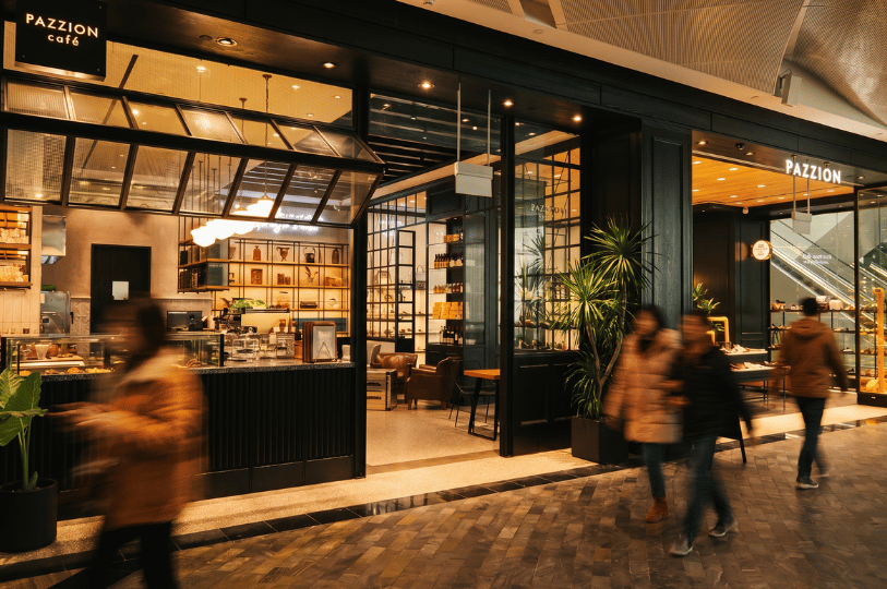 Blurred figures walk swiftly past the storefront of a Café, a hybrid space seamlessly combining a warmly lit coffee shop with a retail shoe store.